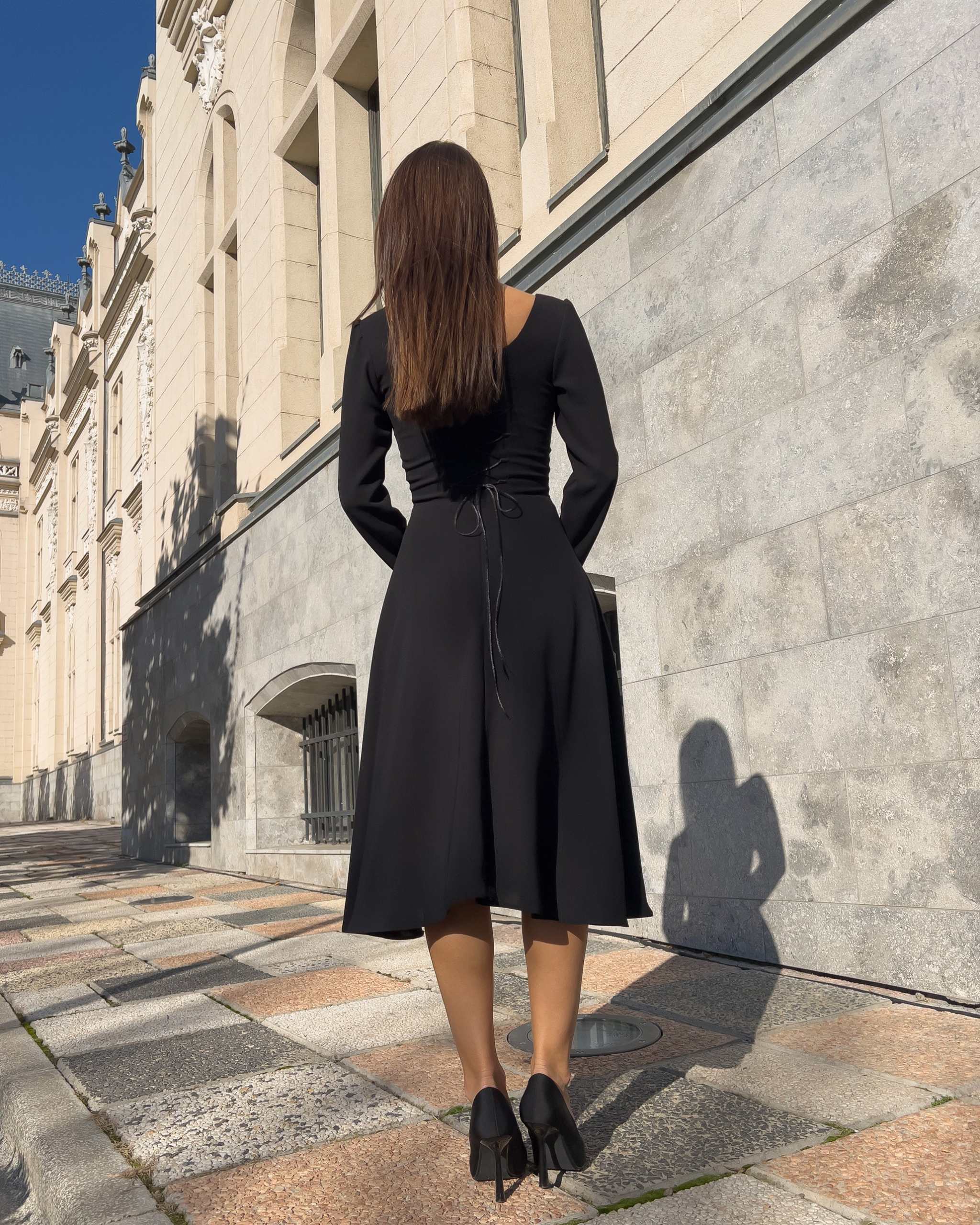 Rochie neagra midi cu mânecă lungă, elegantă, șnur reglabil, și fermoar la spate, în fața unei clădiri.