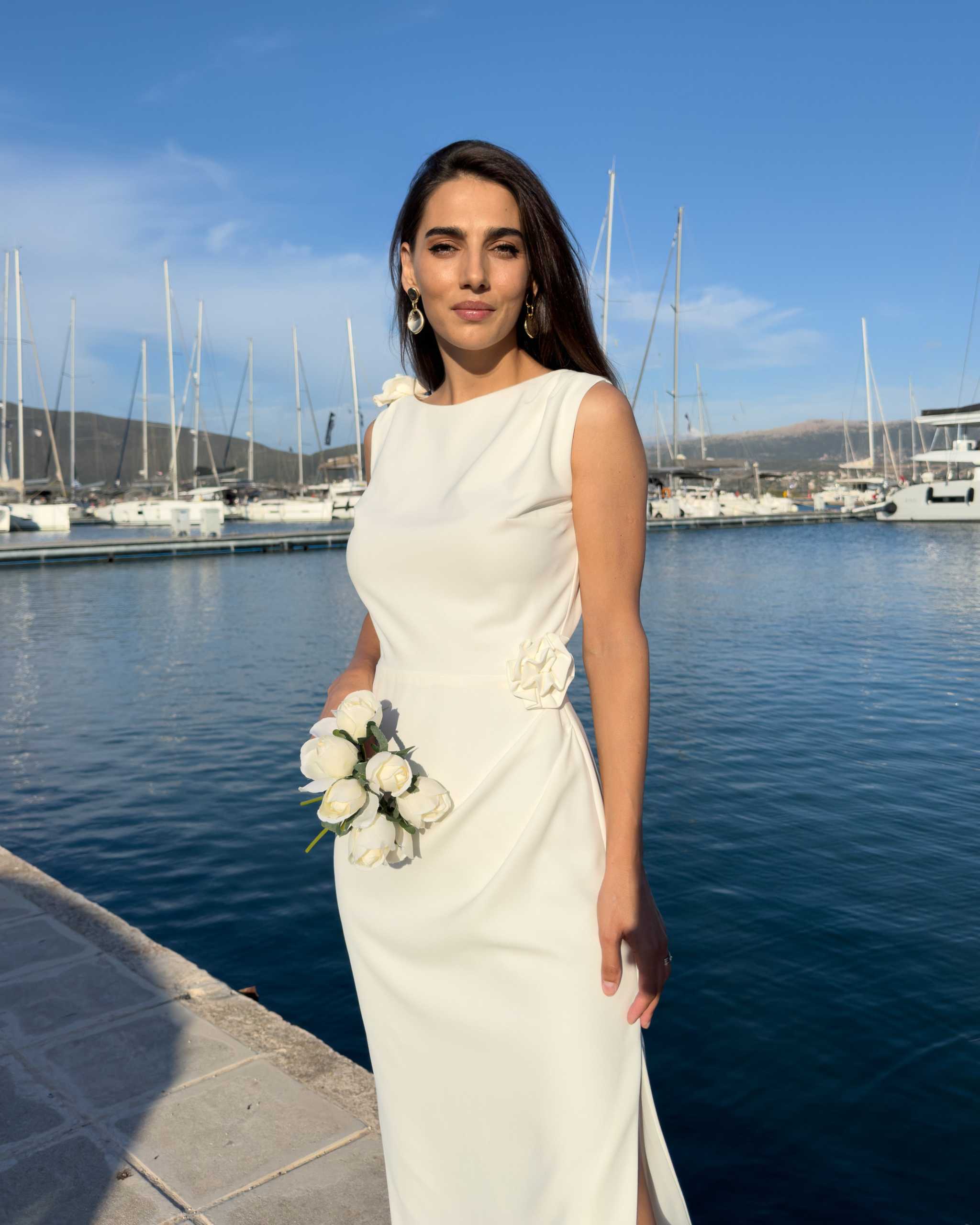Rochie alba de petrecere fără mâneci cu slit și detalii florale, purtată lângă marină, perfectă pentru ocazii formale.