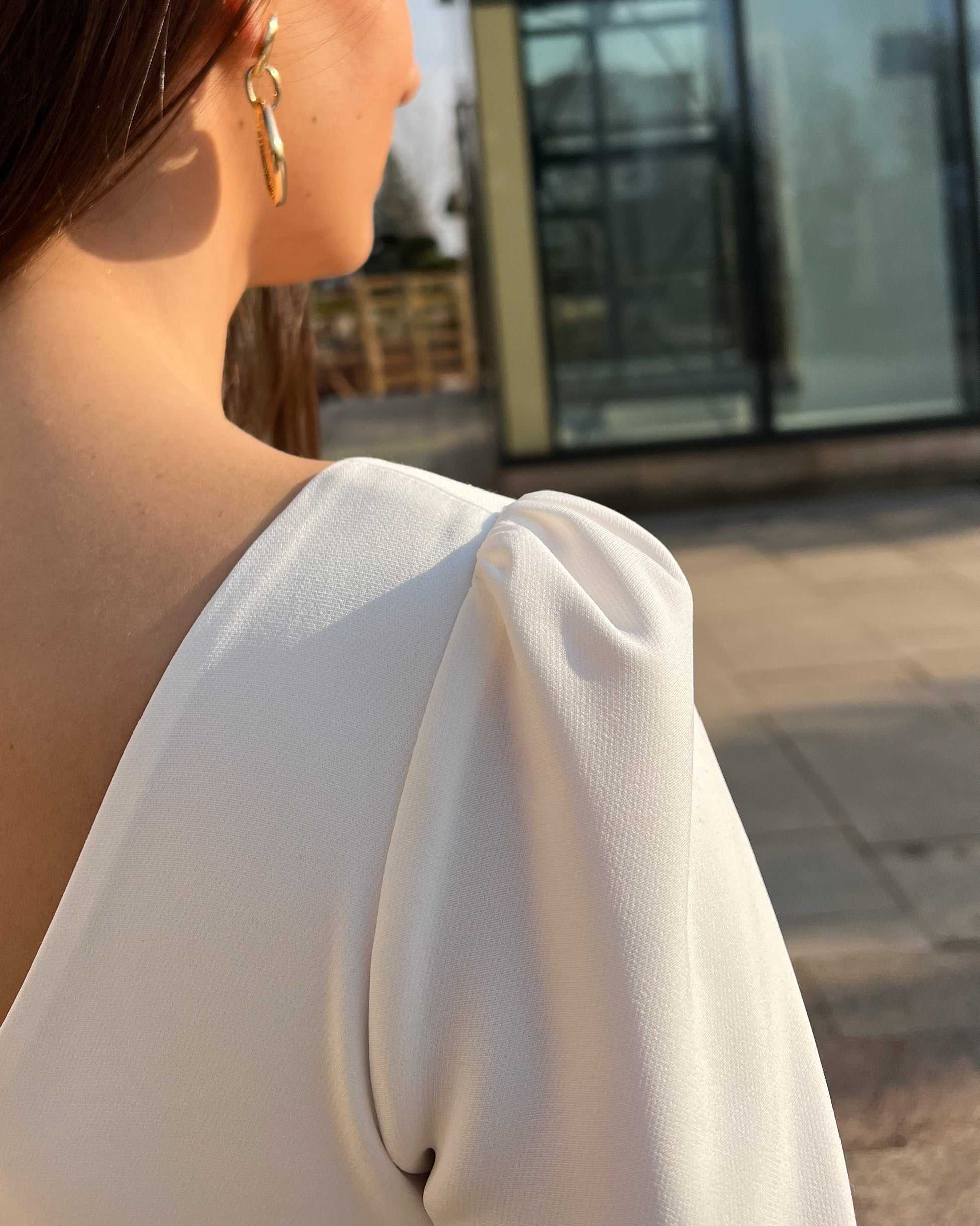 Rochie albă elegantă cu mânecă lungă și detaliu pe umăr, fundal urban.