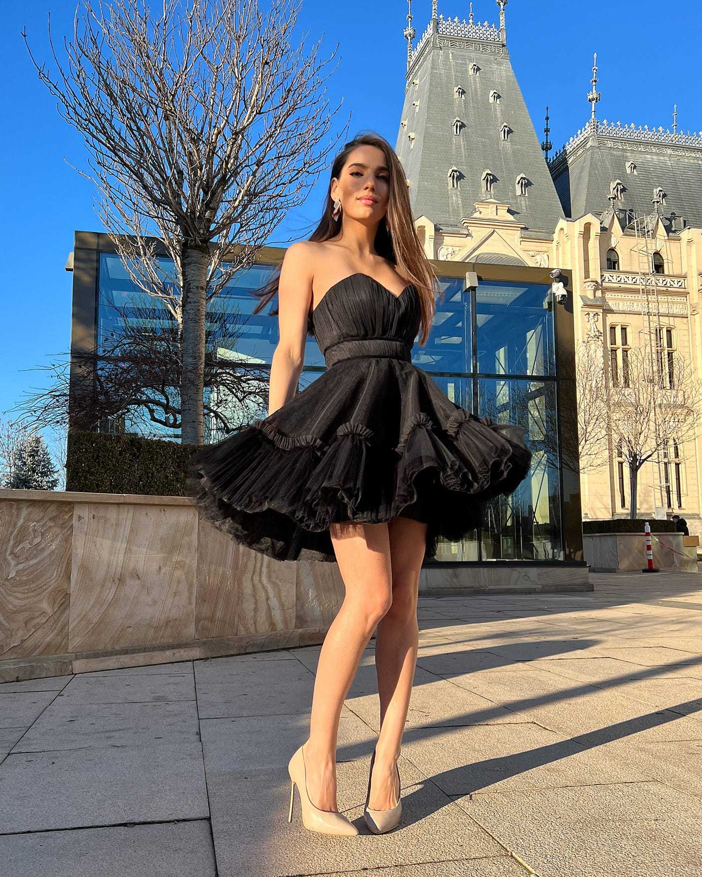 Rochie cocktail elegantă scurtă cu corset negru și fustă evazată, model purtând tocuri înalte, fundal arhitectural.